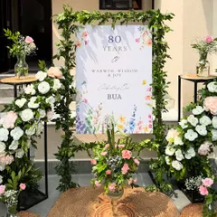 A personalized welcome sign for an 80th birthday, framed with fresh greenery and flanked by beautiful arrangements of pink and white roses. It's the first detail guests see and sets a warm, celebratory tone.