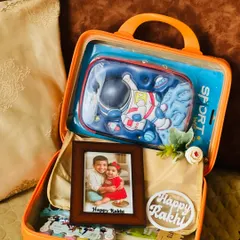 A special Rakhi hamper for a child, packed in a bright orange suitcase. It includes a personalized photo frame and an astronaut-themed pencil case to make the festival extra fun.