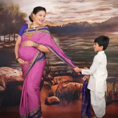 This portrait is inspired by the timeless art of Raja Ravi Varma. The mother-to-be, dressed in a traditional pink and blue saree, shares a gentle moment with her son against a classic, painted backdrop, creating a piece of art that feels both regal and personal.