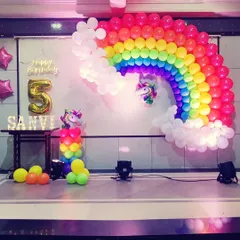 A rainbow and unicorn themed stage decoration for a fifth birthday. A large balloon rainbow arch serves as the main backdrop, accented with a glowing "Happy Birthday" sign, unicorn foils, and a personalized name table for "SANVI".