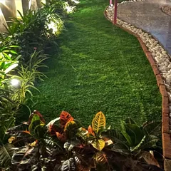 A close-up view of a newly installed artificial lawn at night. The turf is bordered by a brick edge, white pebbles, and a variety of illuminated foliage plants like crotons.