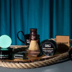A collection of Pink Woolf men's grooming products styled on a rustic rope tray against a dark, moody background. This composition gives the brand a masculine and sophisticated aesthetic.