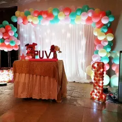 A vibrant and colorful first birthday party for Puvi. This setup includes a multi-color pastel balloon arch over a fairy-light curtain, a personalized name sign on the cake table, and a custom light-up number '1' to mark the special occasion.