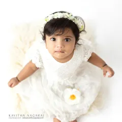 A sweet six-month-old girl in a simple white dress, looking up with wide, curious eyes. The high-key lighting creates a pure and angelic feel.