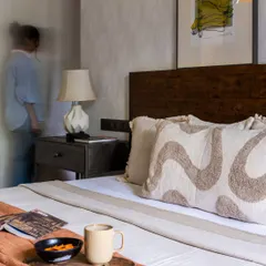 This rustic modern bedroom is infused with a touch of bohemian soul. The motion blur of a person adds a sense of life to the space, which features a dark wood headboard, textured wallpaper, and cozy, patterned cushions.