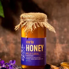 This is a classic product shot for a wild floral honey brand. The rustic jute lid, dark background, and surrounding purple flowers create a moody, organic feel that aligns with the product's natural branding.