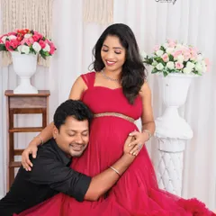 A joyful and candid moment between a couple. The husband-to-be hugs his wife's baby bump as she glows in a vibrant red gown, surrounded by a beautiful floral setup in the studio.