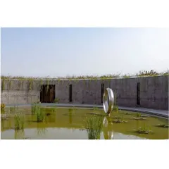 A daytime view of the memorial shows how it blends with the rural landscape. The curved concrete wall encloses a tranquil water court, with a minimalist metallic sculpture at its center. The design creates a sanctuary that feels both grounded and profound. This image also covers media ID 7.