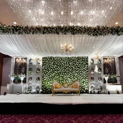 An elegant Walima reception at Crowne Plaza Okhla. The stage features a stunning wall of white flowers, intricate jaali panels, and is set under a magnificent crystal chandelier.