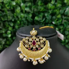 A beautiful chaand bali style maang tikka with a textured gold finish, kundan work, and a central ruby-colored stone.