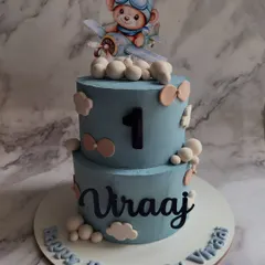 Taking flight for a first birthday! This custom two-tier cake features a hand-modeled pilot teddy bear, fluffy clouds, and personalized details to make a little one's milestone celebration truly special.