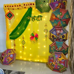 For a traditional Mehendi ceremony, I create a vibrant backdrop using yellow and green fabric drapes, warm fairy lights, and a collection of colorful Rajasthani umbrellas with mirror work. The Hindi "Mehendi" lettering adds an authentic touch.