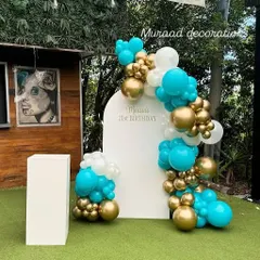 A simple and elegant outdoor backdrop for a 21st birthday. The white arched panel is decorated with a beautiful balloon garland in turquoise, white, and gold, which stands out against the green lawn.