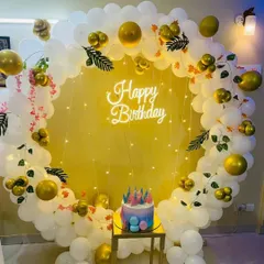 A cheerful birthday photo booth with a circular balloon arch in white and gold. A "Happy Birthday" neon sign and fairy lights add a warm glow, with a small stand ready for the birthday cake.