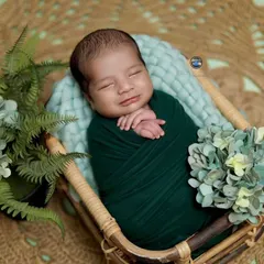 A happy, sleepy smile from this newborn boy, swaddled in a deep forest green wrap. The bamboo basket and surrounding greenery create a natural, earthy vibe.