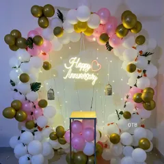 A beautiful and bright anniversary backdrop. This circular frame is decorated with pink, white, and gold balloons, fairy lights, and a glowing "Happy Anniversary" sign, perfect for taking memorable photos.