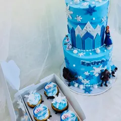 A stunning two-tier Frozen theme cake perfect for a winter princess. The cake is decorated with snowflakes, an ice castle, and beloved characters, paired with a box of matching themed cupcakes.