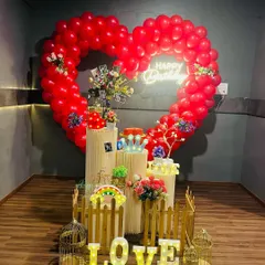 This romantic surprise birthday setup can also be adapted for an anniversary or proposal. It features a large heart-shaped balloon frame, light-up "LOVE" letters, and pedestals for displaying photos and gifts.