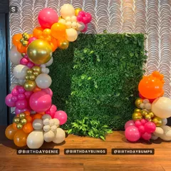A vibrant balloon garland in pink, orange, and gold, set against a lush green hedge wall for a beautiful contrast.