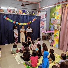A wide shot of the storytelling session, showing the young readers on their 'stage' and the audience of little ones sitting attentively on the floor. This program fosters leadership and mentorship.
