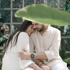 A tender moment captured in a sun-drenched greenhouse. The soft light and abundance of plants create a serene and intimate setting, with the large leaf providing beautiful natural framing.