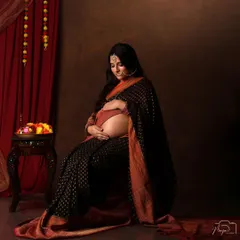 A portrait of serene motherhood. Draped in a stunning black and orange saree, this mom-to-be cradles her bare bump in a classic, seated pose that exudes timeless elegance.