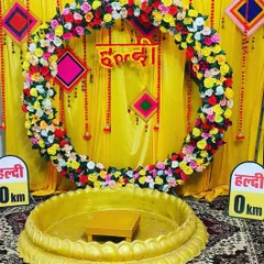 This fun and colorful Haldi backdrop features a large ring frame decorated with multicolored artificial flowers. The setup includes a traditional 'urlī' for the ceremony and quirky 'Haldi 0 km' signs for a playful touch.