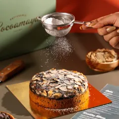 A hand dusting powdered sugar over an almond cake. The falling sugar adds a touch of magic and motion to the photograph.