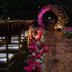 An enchanting evening ceremony setup. The aisle is flanked by vibrant pink lilies leading to a circular arch of flowers and pampas grass, all beautifully illuminated for a romantic nighttime event.