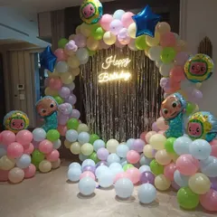 A colorful Cocomelon-themed birthday decoration. I create a circular balloon arch in pastel colors, with a silver tinsel curtain, a 'Happy Birthday' neon sign, and Cocomelon character balloons.
