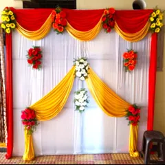 A simple flower and drape decoration for an engagement ceremony at home. The backdrop uses red and yellow fabric with small floral bouquets.