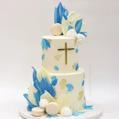 A two-tier baptism cake in white and blue, decorated with abstract chocolate sails, macarons, and a simple gold cross.