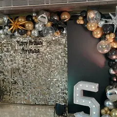 A glitzy birthday setup featuring a silver sequin wall, a balloon arch in black, gold, and silver, and a large light-up number '5'.