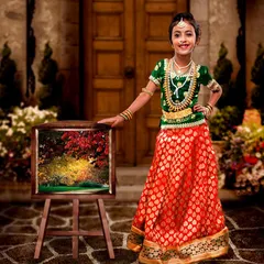 Dressed in a gorgeous green and red lehenga, this young girl poses proudly next to a painting. This setup combines traditional attire with an artistic, classic theme.