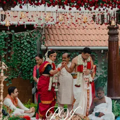 A candid moment from a traditional South Indian Kalyanam. The decor features a canopy of red roses and hanging temple bells, creating an authentic and sacred atmosphere for the rituals.