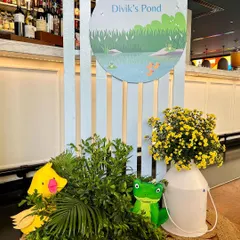 A cute welcome vignette for Divik's duck-themed birthday. The sign says 'Waddle On Into Divik's Pond' and is paired with plush duck and frog toys and fresh yellow flowers.