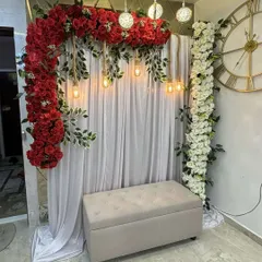 A beautiful and intimate engagement backdrop for a home celebration. It features a stunning arch of red roses, hanging industrial bulbs, and elegant white drapes.