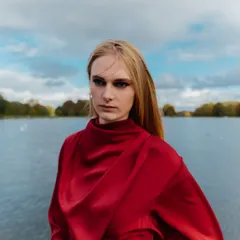 A portrait from an outdoor London shoot. The dramatic sky and open water provide a beautiful, natural backdrop that complements the model's intense gaze and the flowing red top.