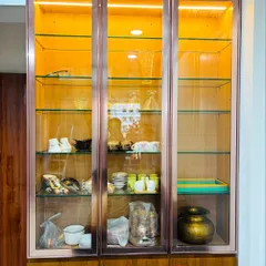 An elegant, freestanding crockery unit designed to showcase your finest dinnerware. This piece features a wooden base with three glass-paneled doors, a rose-gold metallic frame, and warm interior lighting.