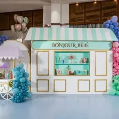 An elegant Parisian-themed baby shower setup, showcasing a custom "Bonjour Bébé" patisserie-style backdrop in mint green and white with gold accents. The display includes a charming Ladurée-inspired cart and is flanked by colorful balloon clusters in blue, pink, and green, creating a chic and welcoming atmosphere.