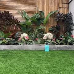A playful and green backyard design. We used high-quality artificial grass for a maintenance-free lawn and created a raised planter bed with a bamboo backdrop, filled with a mix of tropical foliage and decorative statues.