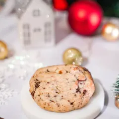 A close-up of a chunky, gooey Christmas cookie on a white pedestal. The simple, clean styling with festive background blur makes the cookie the hero.