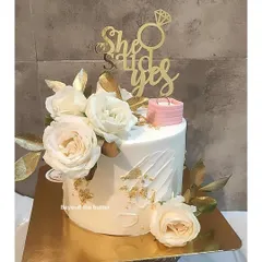 A classic "She Said Yes" engagement cake. The clean white design is elevated with gold-painted leaves, fresh white roses, and a miniature ring box, making it perfect for celebrating the proposal.