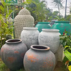 A beautiful assortment of our terracotta and ceramic pots. We offer a range of sizes and finishes, from rustic greys and earthy browns to vibrant sea greens, with a Buddha head statue in the background.