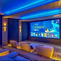 This contemporary media room is designed for both family enjoyment and critical viewing, featuring modern lounge seating with integrated floor lighting. The large-format screen is complemented by blue ambient cove lighting, creating an inviting atmosphere while ensuring excellent picture quality for everything from animated films to blockbuster movies.