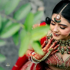 A close-up portrait of the bride, her gentle expression framed by leaves. This shot captures her natural beauty and the richness of her traditional red lehenga.