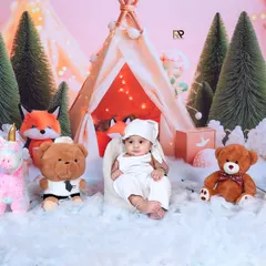 A baby rests in a beanbag chair, surrounded by teddy bears, a unicorn, and a fox in a snowy woodland camp. The warm glow from the teepee creates a cozy and magical winter atmosphere.
