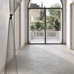 The tall, elegant form of the Ikonika floor lamp in a covered hallway, looking out onto a balcony. This piece is part of the "Collection of Crossed Destinies" and is suitable for both indoor and outdoor use.