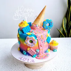 A confectionery-themed cake with a rainbow buttercream base. This design features a pink ganache drip, an upside-down ice cream cone, and lots of sweet treats.
