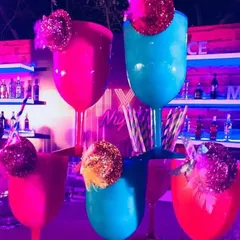 Oversized, brightly colored goblets ready for some disco-era cocktails. The pink and blue glasses, garnished with glittery disco balls, stand out against the neon-lit bar backdrop.
